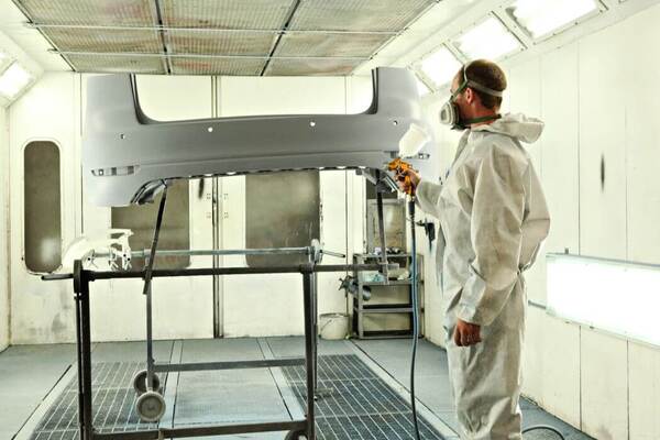 Autolackierer lackiert Stoßstange in moderner Werkstatt, professionelle Fahrzeuglackierung.