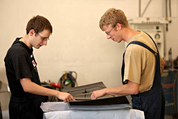 Zwei Auszubildende im Handwerk prüfen gemeinsam ein Metallteil in einer Werkstatt.